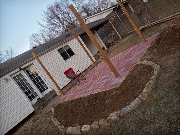 A red patio with wooden posts in a backyard, bordered by a small rock wall and a house with white siding.