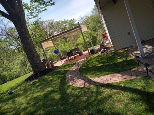 A backyard scene featuring a brick pathway leading to a patio with a table and chairs, with trees and greenery in the background.