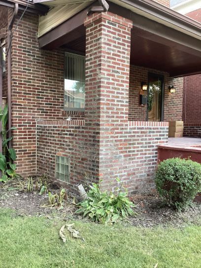 A brick house with a porch, and a small bush in front. A window and an enclosed brick portion are visible.