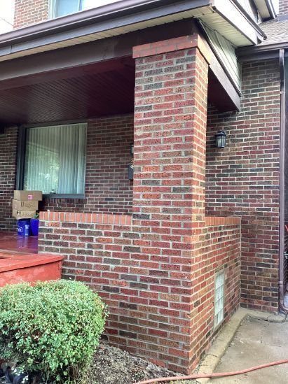 Brick house exterior with a porch supported by a brick pillar, red trim, and a small bush in the foreground.