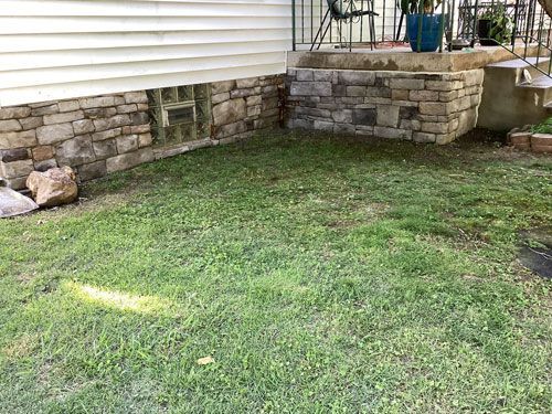 A low stone foundation and porch with a grassy yard. The house is white with a stone facade.