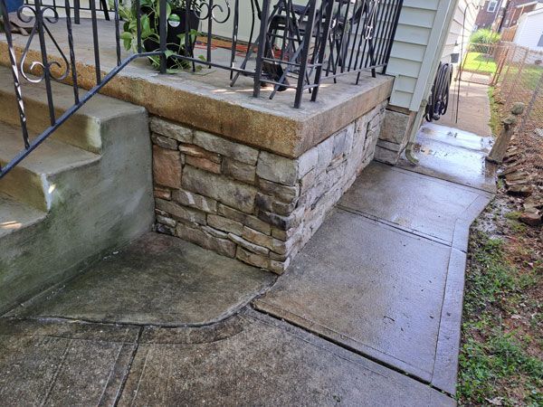 A concrete porch with stone veneer base, wrought iron railing, and a sloped walkway. The concrete is wet.