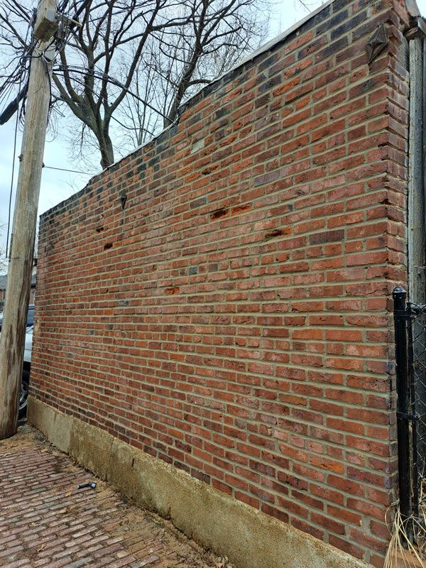 Brick wall with weathered red bricks and a concrete base, leaning slightly. A utility pole and black fence are visible nearby.