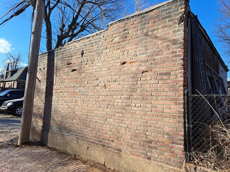 Brick wall of a building, adjacent to a power pole and chain-link fence, with visible damage and a clear blue sky.