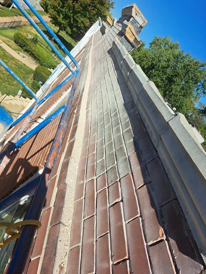 A side view of a building with a brick facade. Blue scaffolding and greenery are visible alongside the building.