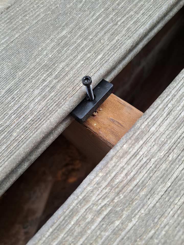 Close-up of composite decking with a hidden fastener clip secured to a wood joist by a black screw.