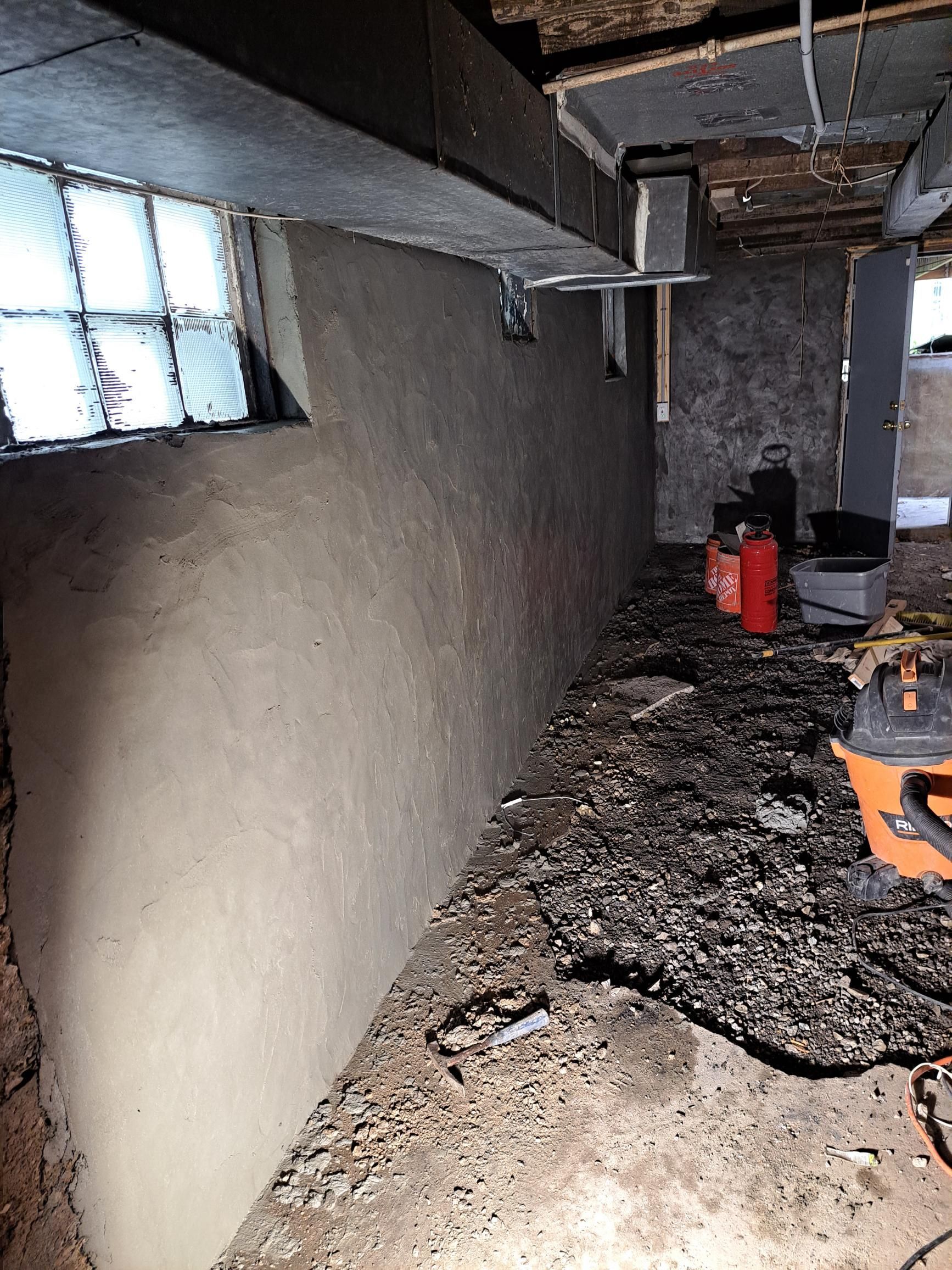 Basement wall coated in light brown plaster, with window and debris on the floor.