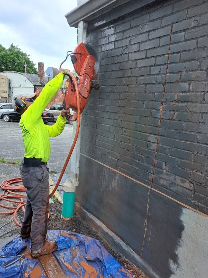 A worker in neon green shirt uses a saw to cut into a dark brick wall, likely for construction. A water hose sprays the cutting area.