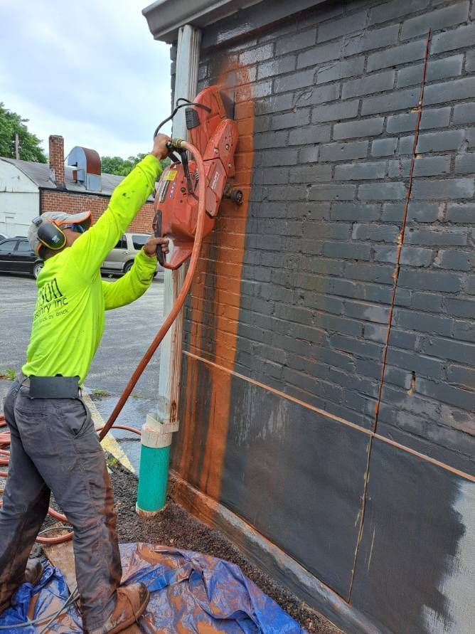 Man in safety gear uses a large saw to cut a vertical line in a brick building exterior. A tarp and water hose are at the base.