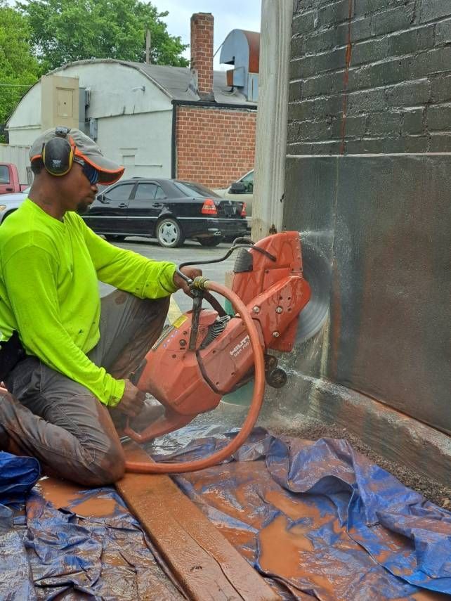 Man in safety gear uses a red power saw to cut into a dark wall. Outdoors.