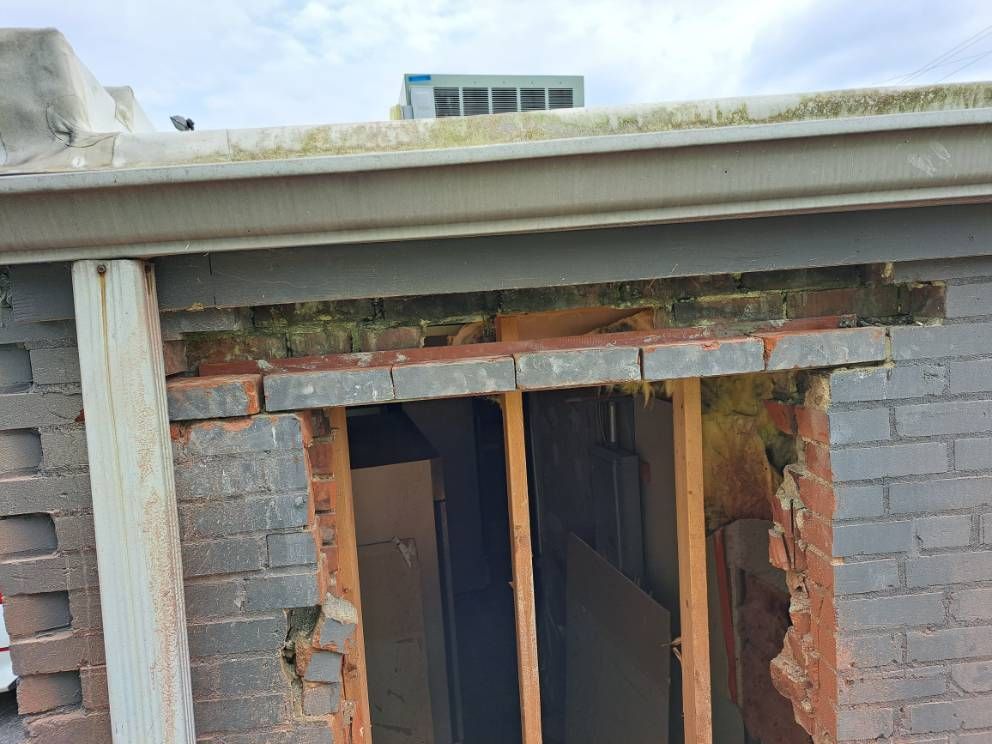 Damaged brick wall with a hole revealing wooden framing, part of a building exterior. Moss grows near the gutter above.