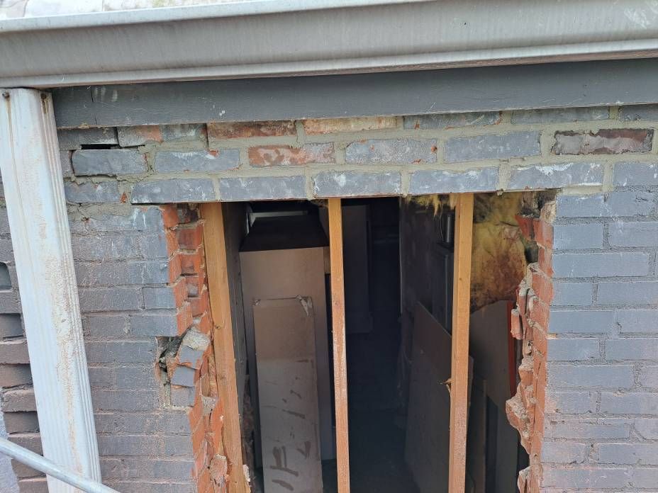 Brick wall partially collapsed with wooden framing inside, revealing a dark interior.