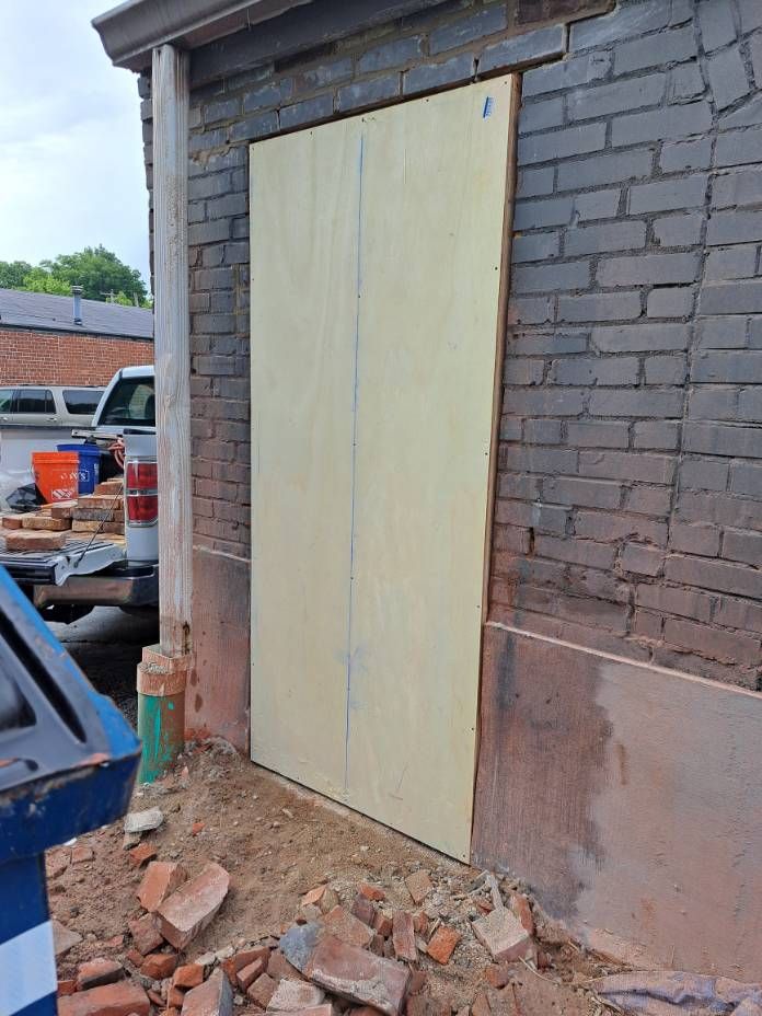 A plywood sheet covers a doorway in a weathered brick building. Construction materials and a truck are visible nearby.