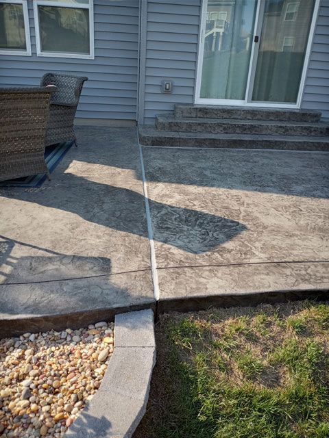 Concrete patio next to a house, with steps leading to a sliding glass door. A stone border and gravel garden sit beside a patch of grass.
