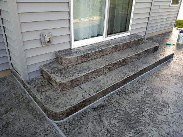 Concrete steps leading to a sliding glass door, textured with a dark grey and brown mottled pattern. Exterior of a building with light grey siding.