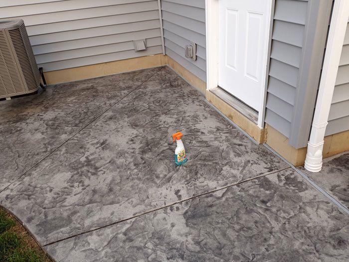 Gray stamped concrete patio next to a light gray house with a white door. An orange and green spray bottle sits on the patio.