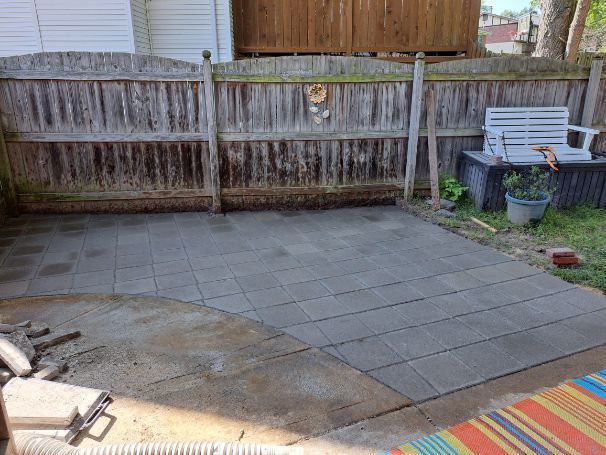 A paved patio in a backyard with a wooden fence and a bench. Concrete pavers fill the space.