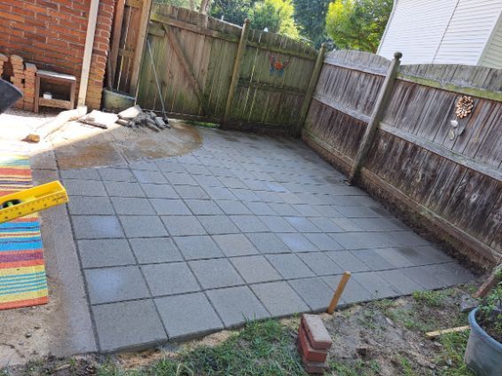 A backyard patio under construction, featuring a grid of gray pavers, bordered by a wooden fence and lush green grass.