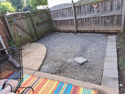Backyard patio with gray gravel, concrete edging, and wooden fences. A circular concrete pad and colorful rug are in the foreground.