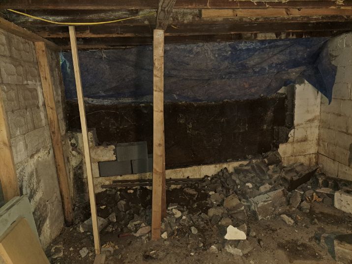 Basement wall demolition in progress; rubble on the floor, supported by wooden posts, tarp covering the top.