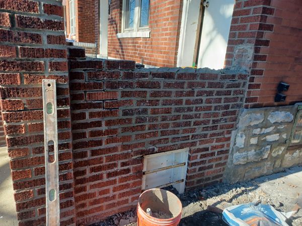 Brick wall construction in progress outdoors; red bricks, concrete base, tools visible.