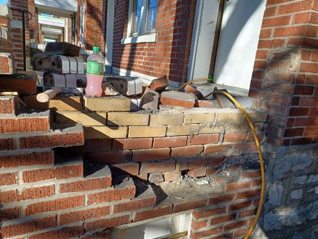 Damaged brickwork on a building's exterior, with exposed yellow brick and loose red bricks. A water bottle and small hammer rest on the brick ledge.