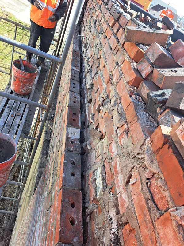 A close-up view of a brick wall being repaired by a construction worker. A narrow gap reveals new brickwork, while old bricks are piled on the roof.