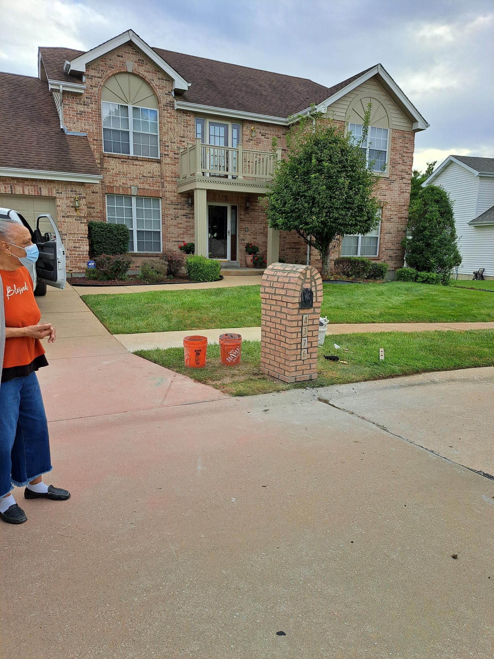 Woman stands on a driveway in front of a two-story brick house. Two orange buckets sit near a brick mailbox.