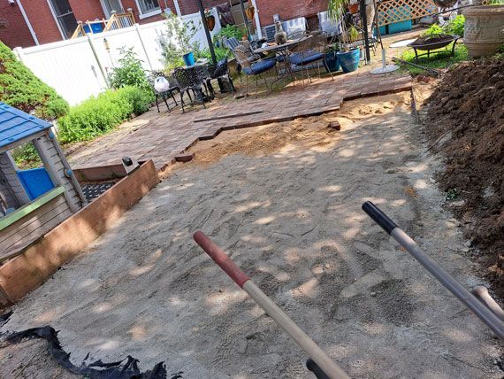 Backyard construction in progress; sand covers an area with partially laid brick paving. Shovels rest on the sand.