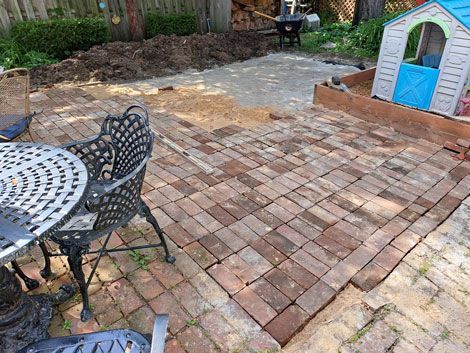 Brick patio under construction, next to a small playhouse. Wrought-iron table and chairs in the foreground; unfinished ground in the background.