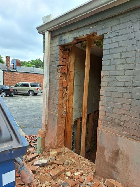 A damaged brick building with a collapsed doorway, bricks scattered on the ground. A dumpster and parked cars are visible in the background.