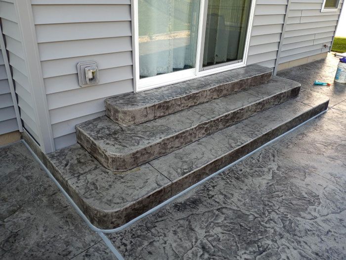 Three concrete steps leading up to a sliding glass door on a gray-sided house. The steps and surrounding patio have a textured, stamped finish.