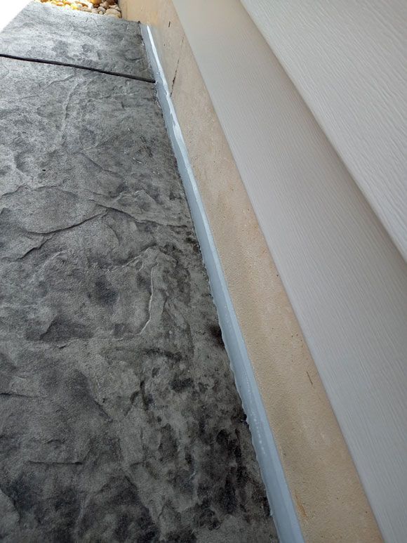 Gray stamped concrete meets a light-colored wall. A thin strip of sealant runs between them.