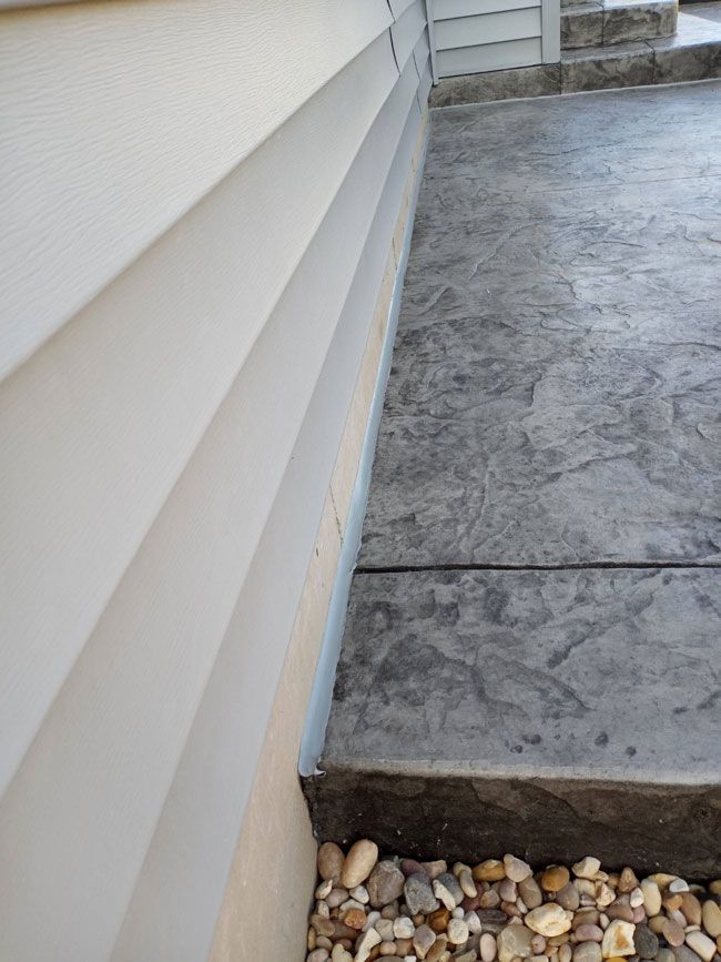 Close-up of a light-colored house siding next to a concrete walkway. Small rocks sit at the base of the wall.