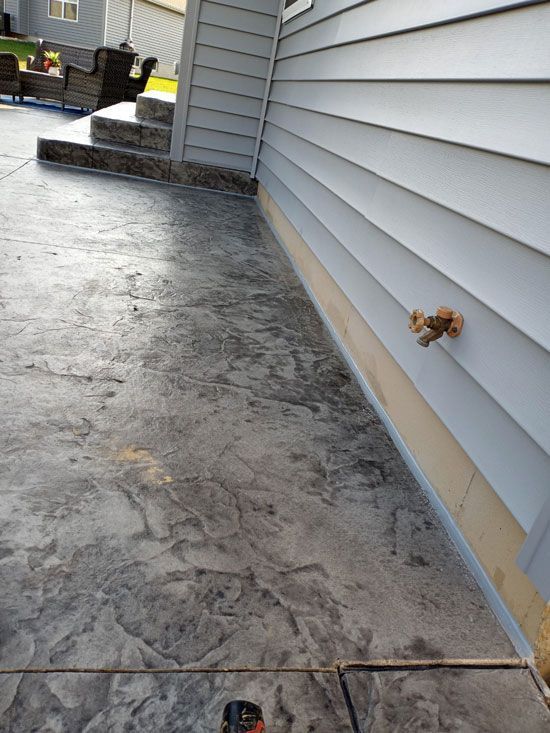 Stamped concrete patio next to a house with light gray siding. The patio has a textured, stone-like appearance.