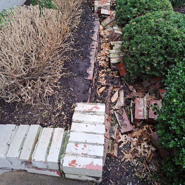 Broken brick edging along a flower bed; some bricks are fallen or dislodged. The bed contains dark soil and bushes.
