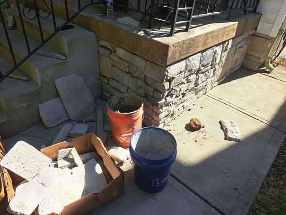 Construction materials on a porch with stone veneer in progress. An orange and a blue bucket sit nearby.