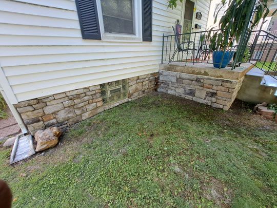 A house with stone veneer along the foundation. The front lawn is green and a small porch with a metal railing is visible.