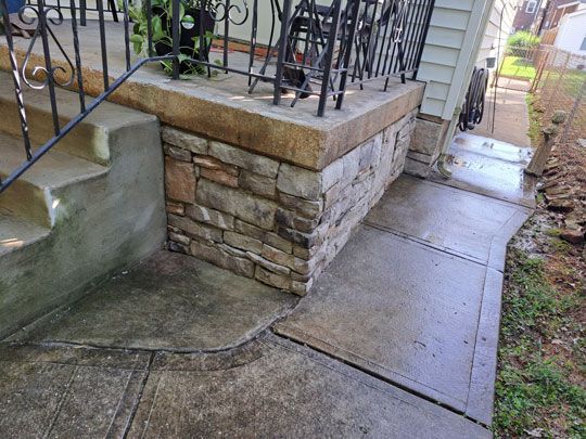 Stone-faced porch and concrete walkway. The walkway leads to the porch steps. The porch has a black railing.