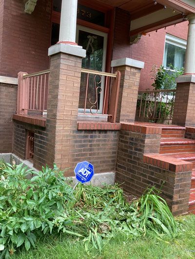 Brick porch entrance of a house with a security sign. Wooden and metal railings, steps, and a doorway are visible. Plants are in front.