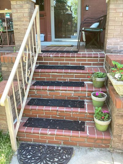 Brick front steps with black mats, a white railing, and potted plants lead up to a glass front door.