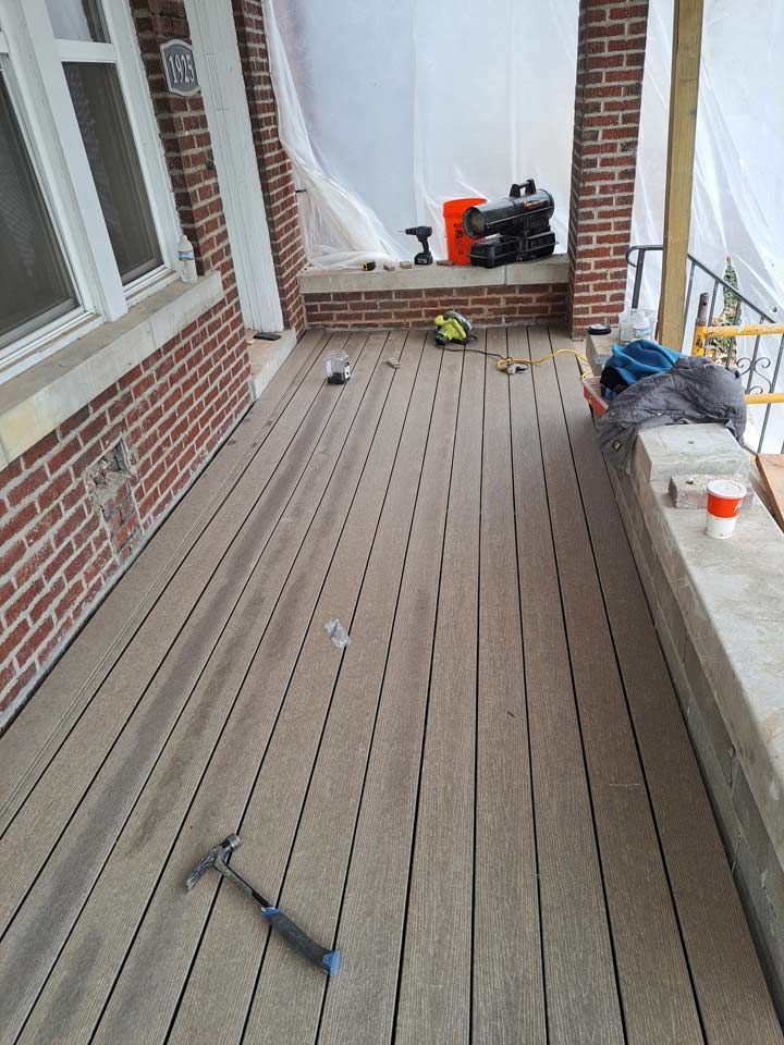 A wooden deck under construction on a house's porch, with tools and supplies scattered around. Brick columns and walls are visible.