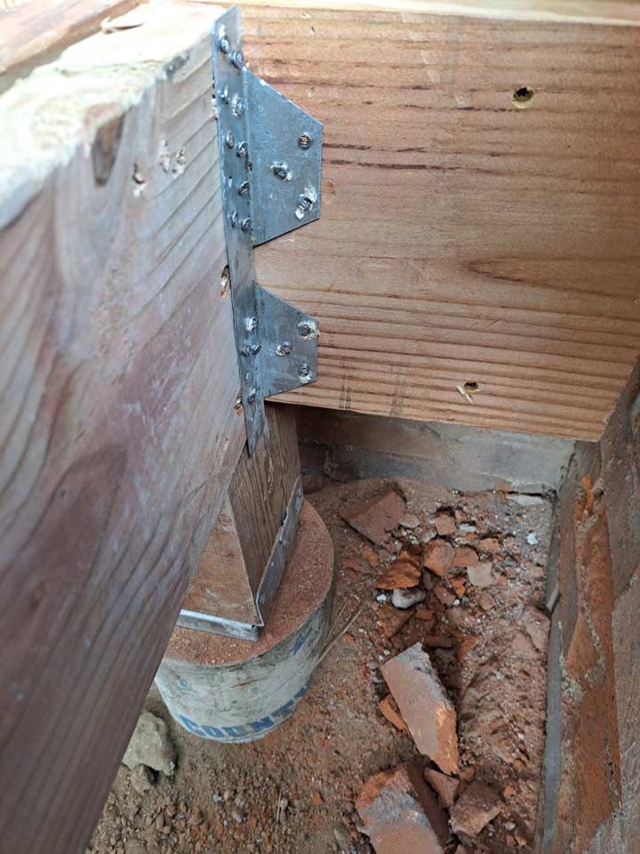 A deck support beam attached to a wooden joist with a metal bracket, resting on a round concrete pier in an outdoor setting with broken bricks.