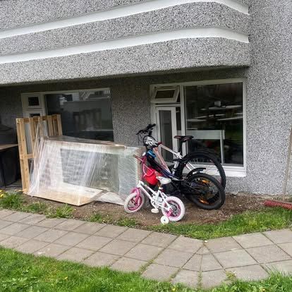 Grátt að utan með gluggum, tveimur reiðhjólum og innpökkuðum viðarkassa á grasinu.