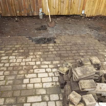 Múrsteinsverönd í byggingu, með hrúgu af múrsteinum, skóflu og mold upp við trégirðingu.