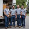 Four men in gray shirts and blue jeans stand in front of a moving truck.