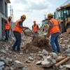 Construction workers in orange vests and hard hats removing debris from a road. Yellow heavy equipment visible.