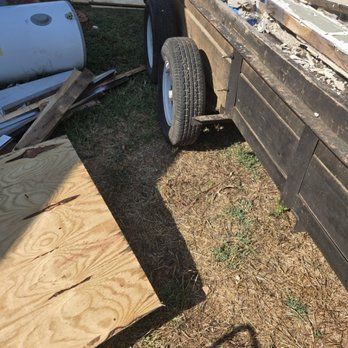 A trailer loaded with debris, including wood and sheet metal, sits on grass, near a white tank.