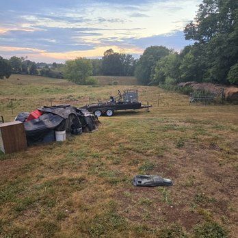 Grassy field with hay bales, a trailer, and assorted equipment under a cloudy sky.