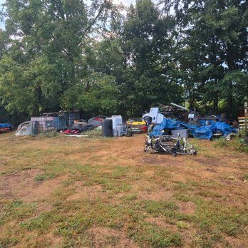A cluttered outdoor area with scrap metal, car parts, and tarps, set against trees and a grassy field.